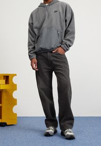 Šedá mikina s kapucí a přední kapsou; v kombinaci s tmavě šedými širokými kalhotami a šedými teniskami s černými akcenty. V pozadí žlutý nábytek.