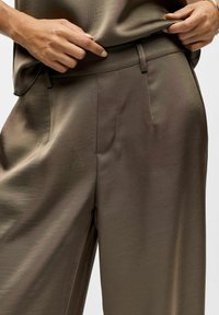 Hände passen den Bund von glänzenden braunen Satin-Hosen an, die mit einem passenden Oberteil kombiniert sind, gezeigt von der Taille bis zur Mitte des Oberschenkels.