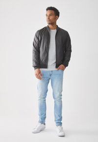 Sort bomberjakke, grå sweater, lyseblå jeans og hvide sneakers. Jakken har en lynlåslukning foran og ribbede ærmer.