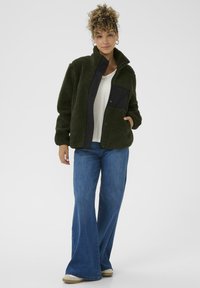 Vachtige donkergroene jas met een zwarte ritszak, hoge kraag en lange mouwen, gecombineerd met een lichtgekleurde shirt en blauwe flared jeans.
