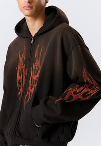 Sweat à capuche noir zippé avec des motifs de flammes rouges et orange sur le devant et les manches. Confectionné en tissu doux avec une coupe décontractée et une capuche.
