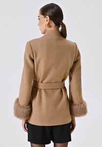 Cappotto color cammello con finitura testurizzata, caratterizzato da maniche lunghe con polsini in pelliccia sintetica e una vita abbinata con cintura. Vista posteriore mostrata.