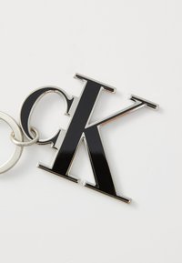 Porte-clés en métal noir présentant une lettre "K" surdimensionnée et brillante avec des accents en argent et un anneau d'attache circulaire.