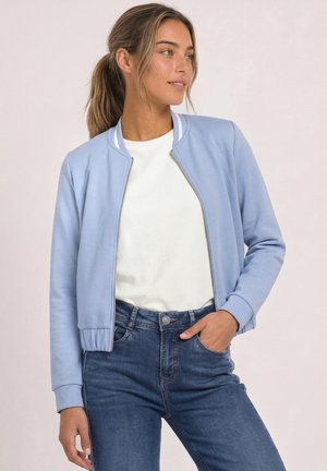 Femme aux cheveux châtain clair attachés en queue de cheval, vêtue d'une veste bomber bleu clair, d'une chemise blanche texturée et d'un jean bleu, regardant vers la droite.
