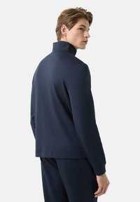 Námořnicky modrý pulovr s vysokým límcem, vyrobený z měkké látky, s volným střihem a dlouhými rukávy. Hladká textura bez viditelných vzorů.