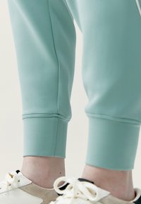 Pantaloni sportivi verde menta con orli alle caviglie, realizzati in tessuto liscio, abbinati a sneaker bianche e beige con lacci e dettagli.