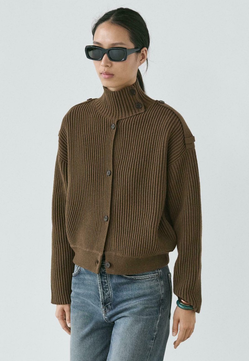 Brauner, gerippter Strickcardigan mit hohem Kragen, Knopfdetails und verkürztem Schnitt, kombiniert mit blauen Jeans. Glatte Textur und lockere Silhouette.