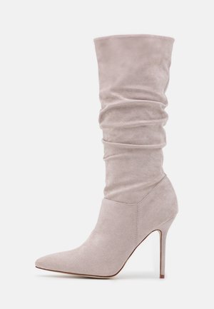 Botas de ante beige claro hasta la rodilla con punta afilada y tacón de aguja, que presentan un diseño arrugado en el eje.