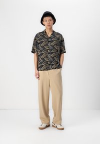 Camisa negra de manga corta con patrón de hojas doradas, pantalones de pierna ancha beige, sombrero negro y zapatillas blancas con detalles negros.