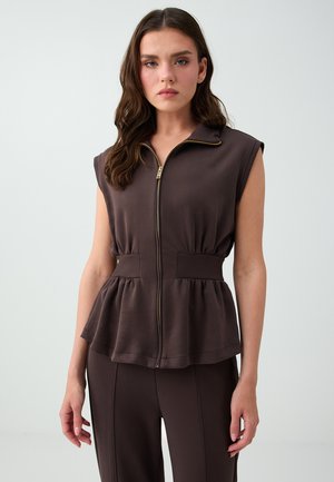 Vrouw met lang bruin haar, gekleed in een mouwloos donkerbruin zip-up topje met een geplooide taille en bijpassende broek tegen een effen achtergrond.