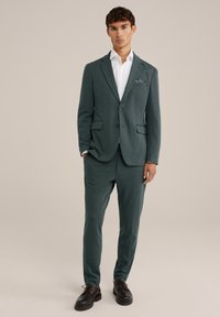 Men's suit in dark green fabric with textured pattern. Features two front pockets, single-button closure, and matching tailored trousers. Black shoes.