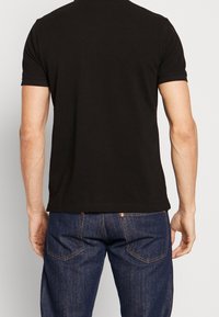 Uomo che indossa una semplice t-shirt nera a maniche corte e jeans di denim blu scuro, visto da dietro su uno sfondo neutro.