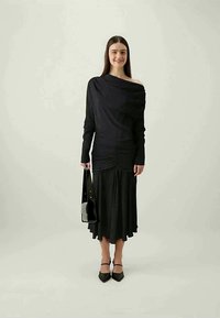 Crna haljina dugih rukava s naboranom teksturom u struku, s lepršavom suknjom, nosi se uz crne štikle i drži pravougaonu crnu torbicu.