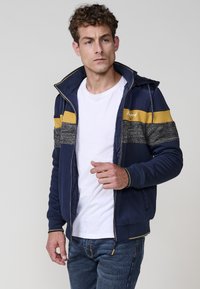 Chaqueta de navy con cremallera, con rayas horizontales amarillas y grises, acentos de tela texturizada y capucha. Camiseta blanca debajo, ajuste casual.