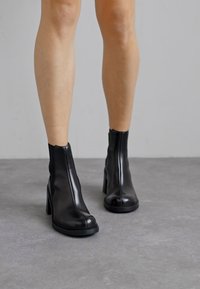 Botas de tobillo de cuero negro con una textura suave, cierre de cremallera y un tacón grueso, que presentan una puntera redonda y un diseño minimalista.