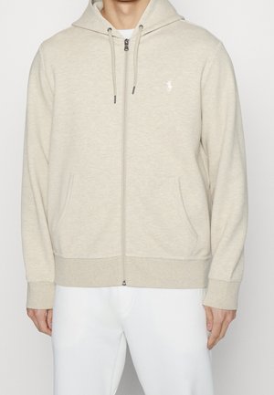 Homme portant un sweat à capuche beige zippé avec poches avant et un petit logo blanc sur la poitrine, associé à un pantalon blanc.