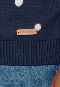 Marineblauer Pullover mit weißen Punkten, geripptem Saum und einem hellbraunen Lederetikett mit der Aufschrift "EDC BRAND" am unteren Rand. Glatte Textur.