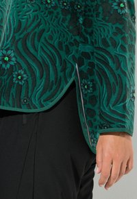 Zielona bluza polarowa z kwiatowymi i abstrakcyjnymi wzorami. Posiada zaokrąglony dół i rozcięcia po bokach, obszyte ciemnozieloną lamówką. Noszona na czarnych spodniach.