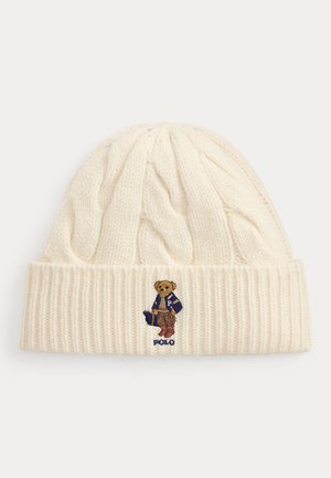 Gorro tejido de color crema con borde doblado que presenta un oso bordado con chaqueta azul y pantalones marrones sosteniendo una bolsa.