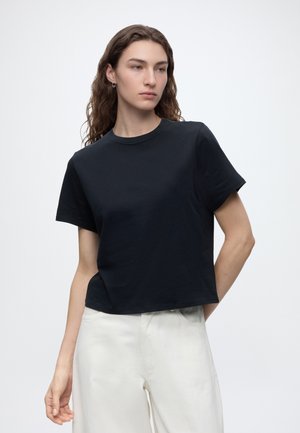Giovane persona con capelli lunghi e ricci che indossa una maglietta nera a maniche corte e pantaloni bianchi, guarda di lato su uno sfondo semplice.