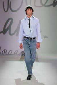 Chemise blanche à manches longues, jeans bleu clair, ceinture noire et chaussures noires. Le modèle porte une casquette grise, mettant en valeur un look décontracté et superposé.