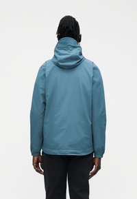 Person with dark skin and dreadlocks wearing a blue hooded jacket and black pants, viewed from the back against a plain white background.