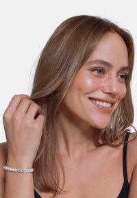 Bracciale di perle bianche con design a catena. Modella che indossa un top nero, sorridente con capelli lunghi e lisci di colore castano e carnagione chiara.