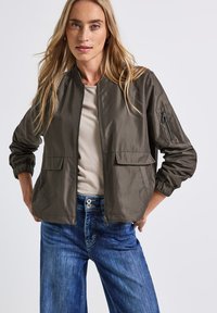Olivgrön bomberjacka i lätt material med framfickor, en zip-ficka på ärmen och elastiska manschetter, kombinerad med blå jeans.