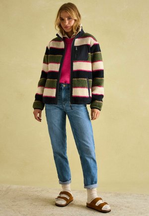 Multicolored fleece jacket with black, cream, green, and pink stripes, a zip pocket, paired with blue jeans and brown sandals.