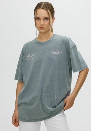 Joven mujer con cabello castaño claro recogido en una coleta, vistiendo una camiseta gris suelta con los textos "Matcha" y "Lovers Club", combinada con pantalones blancos.