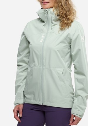 Femme portant une veste zippée vert clair à col haut et un pantalon violet, debout devant un fond blanc.