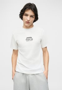 Νέος άνδρας με σκούρα μαλλιά φοράει λευκό T-shirt Replay με μαύρο κείμενο και γκρι φόρμα, στέκεται μπροστά σε ένα απλό φωτεινό φόντο.