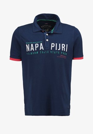 Marineblauwe polo met rode accenten, voorzien van een logo en tekstprint, gecombineerd met lichtbeige jeans en bruine sneakers.