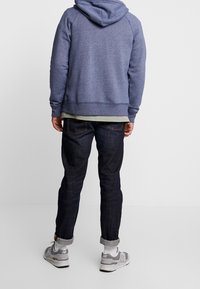 Marinblå huvtröja med snörning, lagd över en ljusgrön skjorta, parat med mörkblå jeans och grå sportskor. Casual passform.