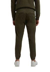 Homme portant un pantalon de jogging vert foncé et des baskets blanches, montré de dos sur un fond blanc.