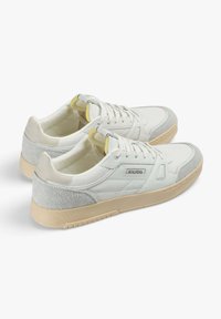 Zapatillas blancas con acentos de ante gris, parte superior de lienzo texturizado, suela de goma beige, punta redonda y cordones. Presenta el logo de la marca en el lateral.