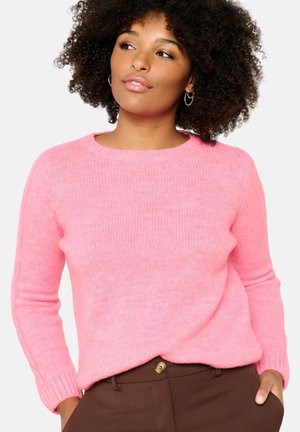 Rosa gestrickter Pullover mit langen Ärmeln und Rundhalsausschnitt, aus weichem Material. Getragen mit braunen Hosen, die einen goldenen Knopf haben.