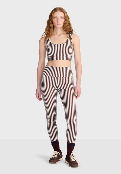 Jeune femme aux cheveux rouges bouclés portant un crop top rayé vertical marron et blanc et des leggings avec des chaussettes foncées et des baskets marron.