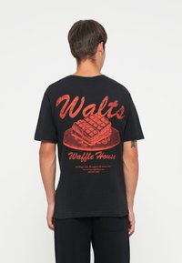 Musta puuvillapaita, jonka selässä on punainen graafinen kuva pöyd�ällä olevasta vohvelista, jossa on teksti "Walt's Waffle House" ja yhteystiedot.