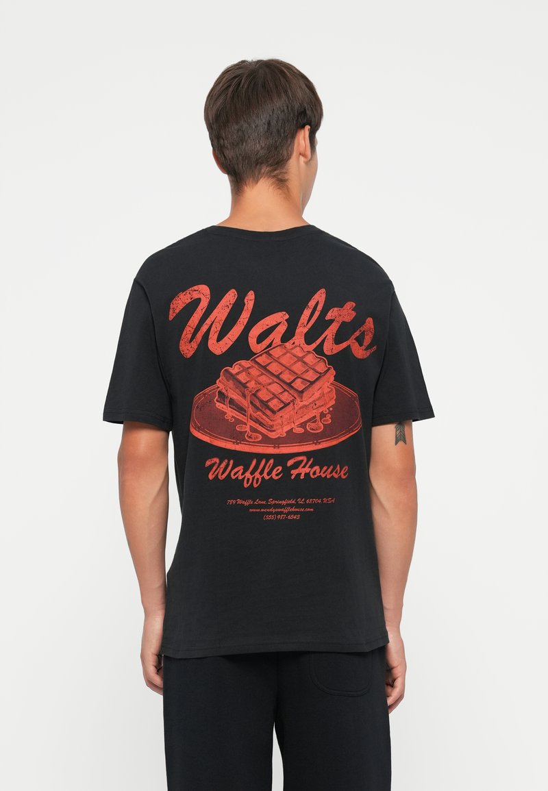Musta puuvillapaita, jonka selässä on punainen graafinen kuva pöydällä olevasta vohvelista, jossa on teksti "Walt's Waffle House" ja yhteystiedot.