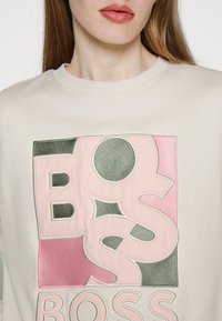 Felpa color beige chiaro con una grande grafica texturizzata in rosa, verde e nero, con la parola "BOSS" ben visibile.