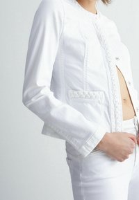 Valkoinen lyhyt denim-takakiinnitys, jossa on palmikkoreunus ja hopeiset napit. Takissa on pitkät hihat ja etutasku, ja se on yhdistetty valkoisiin farkkuihin.