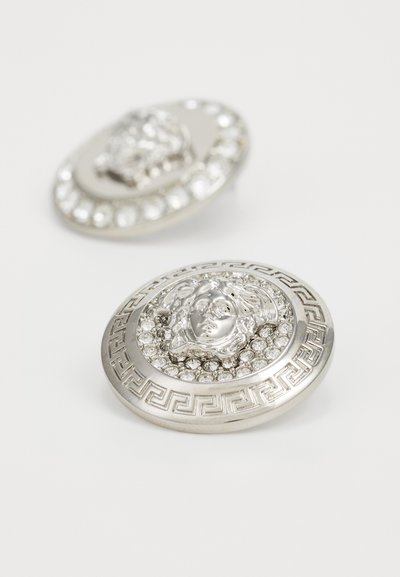 Stříbrné brože s vyvýšeným designem obličeje obklopené kruhem průhledných krystalů a vzorem řeckého klíče na okraji.