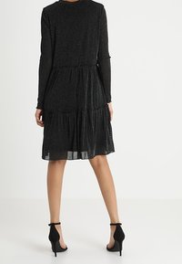 Black long-sleeve dress with layered tiers, made of sparkly fabric, featuring a round neckline and paired with black high-heel shoes.