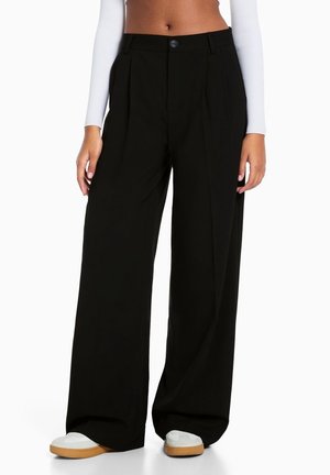 Femme portant un pantalon noir taille haute à jambes larges, associé à des baskets blanches et un haut blanc manches longues court.