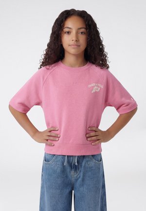 Joven con cabello rizado de pie con las manos en las caderas, vistiendo una camiseta rosa de "Atletismo" y jeans azules sueltos contra un fondo liso.