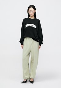 Zwarte sweatshirt met witte tekst, gecombineerd met losse, lichtgroene broek en zwarte schoenen met hak en puntige neus. Draagt een bril met een helder montuur.