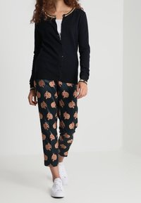 Marineblauer Cardigan mit rundem Halsauschnitt, goldenen Akzenten und Knopfverschluss. Gemusterte Hose in Dunkelblau mit orangen Formen. Weiße Sneakers.