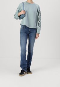 Sweatshirt bleu clair avec détails décoratifs sur les manches, associée à un jean bleu foncé et des baskets noires avec semelles en gomme.