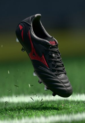 Black soccer cleat with red accents, textured synthetic upper, six white studs, and a laced design, depicted above green grass.